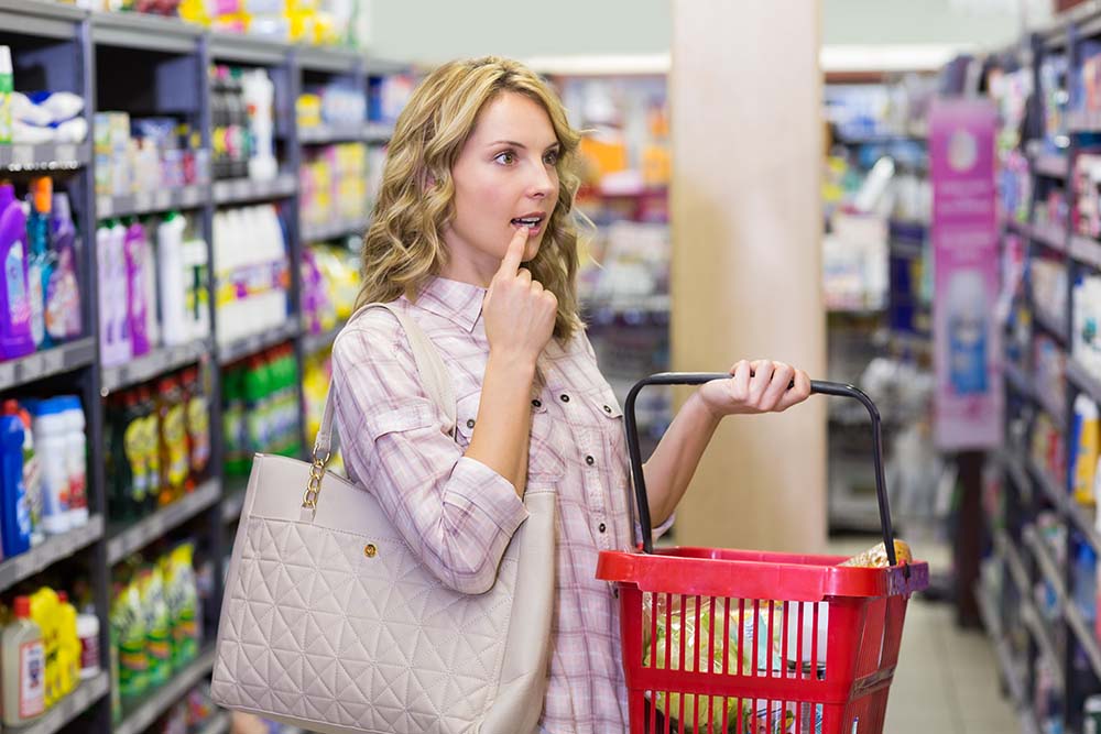 Женщина с сумкой для покупок задумчиво смотрит на полку в супермаркете