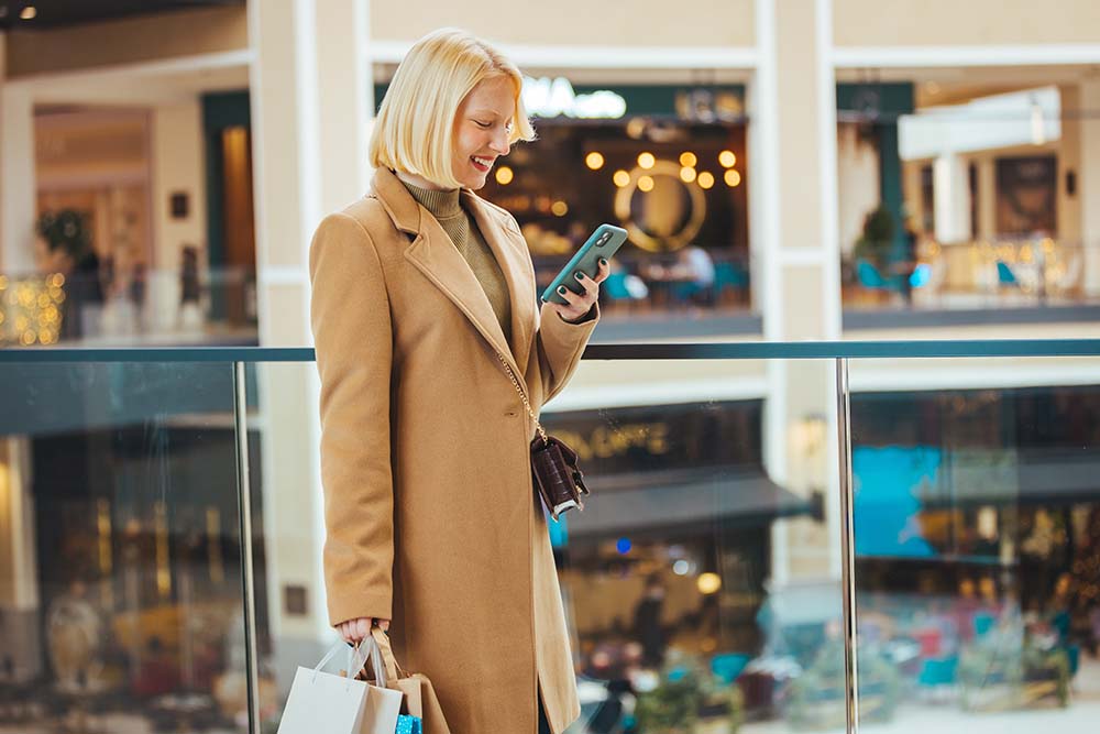 Молодая женщина с сумками для покупок пользуется смартфоном, стоя в магазине (Болгария)